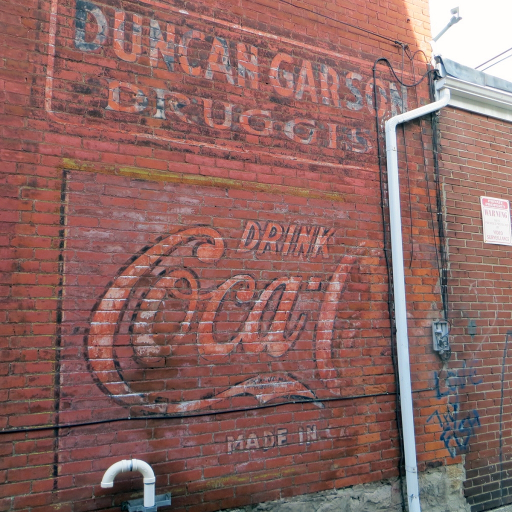 Faded Coca-Cola ad on exterior brick wall below a similarly faded druggists shop name.